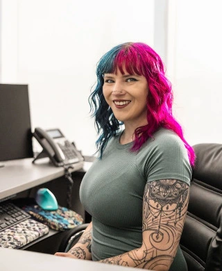 Smiling woman with pink and blue hair, tattoos on arms, seated at a desk with computer and phone.